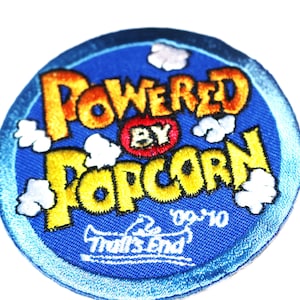 May include: A blue and yellow embroidered patch with the text "Powered by Popcorn" and a trumpet logo with the text "The End" and "09-10".