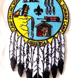May include: Embroidered patch with a circular design featuring a blue background with various illustrations and text. The patch includes a yellow border and a series of white and black feathers hanging below. Text includes "Camp Falling Rock Simon Kenton Council Summer Camp".