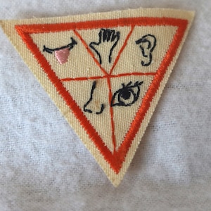 May include: A triangular patch with an orange border and five embroidered images: a closed eye, a nose, a hand, an ear, and a tongue. The images are arranged in a circle around a central point.