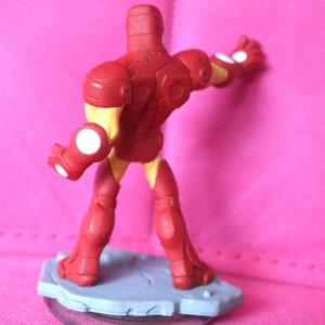 May include: A red and yellow Iron Man action figure, viewed from the back, standing on a gray base. The figure has a detailed design with white accents on the hands. The background is a solid pink colour.
