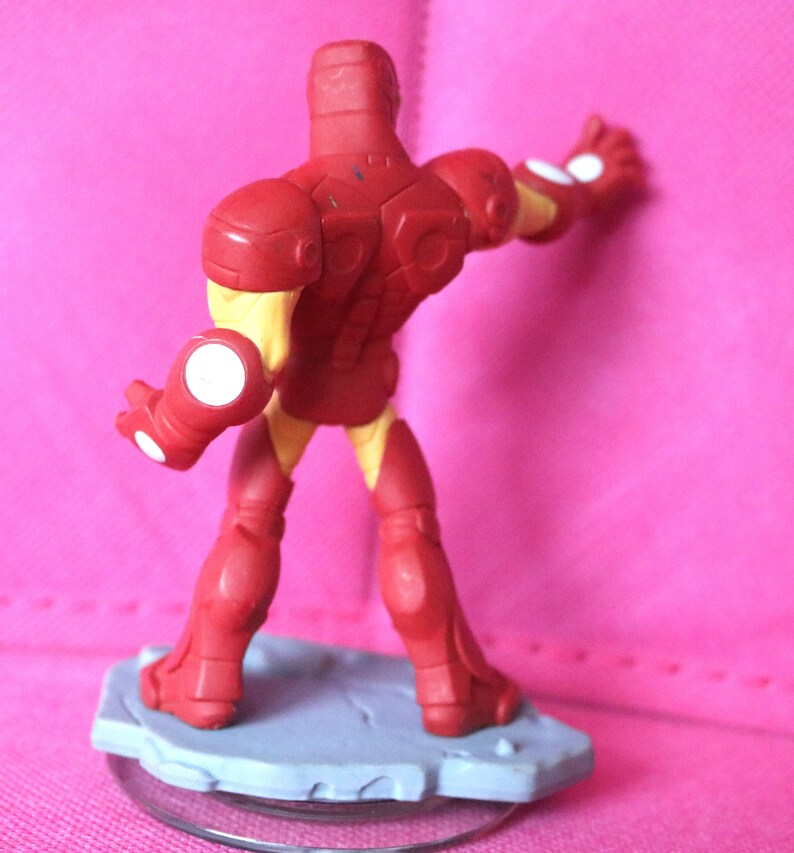 May include: A red and yellow Iron Man action figure, viewed from the back, standing on a gray base. The figure has a detailed design with white accents on the hands. The background is a solid pink colour.