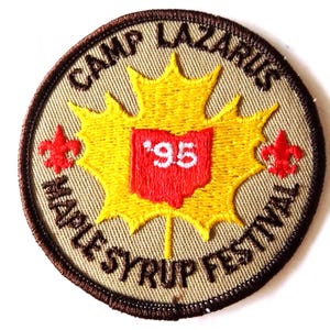 May include: A circular patch with a tan background and a brown border. The patch features a yellow maple leaf with a red Ohio-shaped center containing the number '95'. The text 'CAMP LAZARUS' and 'MAPLE SYRUP FESTIVAL' are embroidered around the edge, along with two red fleur-de-lis symbols.