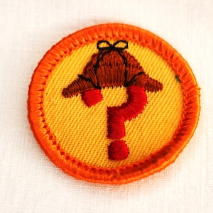 May include: A yellow and orange embroidered patch with a red question mark and a brown hat on top.