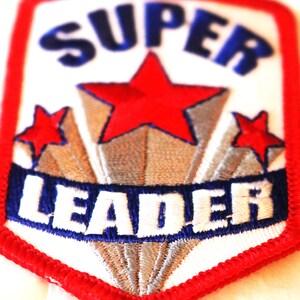 May include: Embroidered patch with a red border and a white background. The patch features the word "SUPER" in blue, a large red star, two smaller red stars, and the word "LEADER" in white on a blue banner.