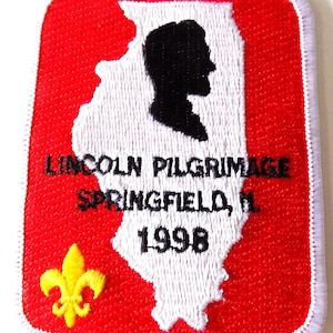 May include: A rectangular patch with a red background and a white border. The patch features a white outline of the state of Illinois, with a black silhouette of Abraham Lincoln. Text reads "LINCOLN PILGRIMAGE SPRINGFIELD, IL. 1998". A yellow fleur-de-lis is in the lower left corner.