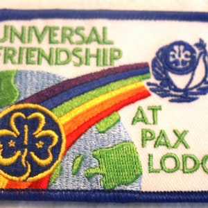 May include: Embroidered patch with a blue border and the words "UNIVERSAL FRIENDSHIP AT PAX LODGE." Features a rainbow, a globe, and a trefoil emblem. The patch is rectangular with rounded corners.
