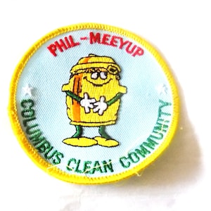 May include: Embroidered patch with a yellow cartoon character wearing sunglasses and a hat. The patch has a light blue background with the words "PHIL-MEEYUP" and "COLUMBUS CLEAN COMMUNITY" in red and green, respectively. A yellow border surrounds the patch.