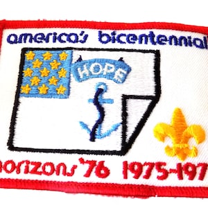 May include: A rectangular embroidered patch with a red border on a white background. It features the text 'america's bicentennial' in blue, a blue flag with yellow stars, a blue anchor, a blue banner reading 'HOPE', and a yellow fleur-de-lis.