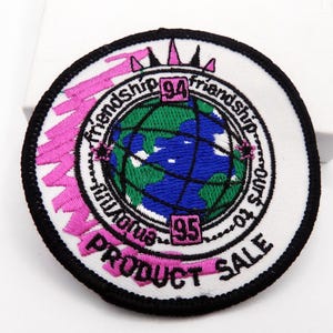 May include: Embroidered patch with a white background, black border, and a globe design. The globe is green and blue, with the text "Friendship" and "Product Sale" in pink. The numbers "94" and "95" are also visible.