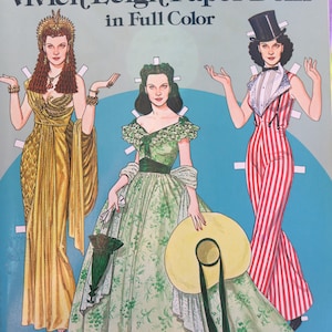 May include: A vintage paper doll book featuring three paper dolls of actress Vivien Leigh. The dolls are dressed in different outfits, including a gold gown, a green dress with a floral pattern, and a red and white striped pantsuit. The book cover features the title "Tom Tierney Vivien Leigh Paper Dolls in Full Color".