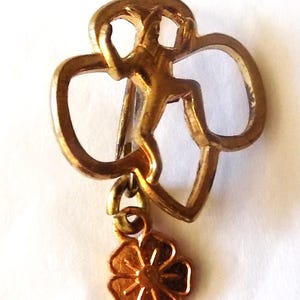 May include: A vintage gold-toned metal pin featuring a stylized figure within a trefoil shape, with a small flower charm dangling below. The pin has a classic design, likely a collectible item.