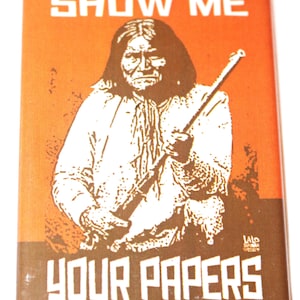 May include: Rectangular button with an orange background and brown text and image. The text reads "SHOW ME" above an illustration of a Native American man holding a rifle. The text "YOUR PAPERS" is below the image.