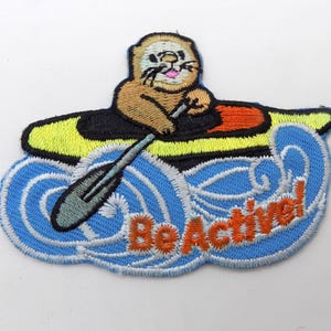 May include: Embroidered patch featuring a cartoon otter in a kayak, paddling through blue waves. The kayak is yellow and black, with an orange accent. The text "Be Active!" is in orange on the waves.