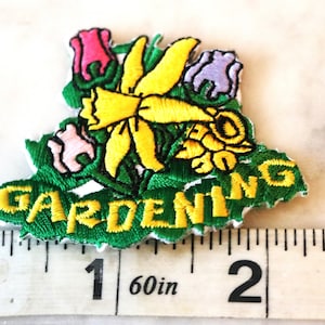 May include: Green felt patch with yellow embroidered text "GARDENING" and a yellow embroidered daffodil with pink and purple flowers. The patch is about 2 inches wide.