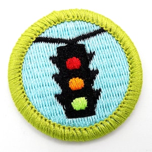 May include: Embroidered patch featuring a black traffic light with red, orange, and green lights. The traffic light is centered on a light blue background, surrounded by a green stitched border. The patch is round.