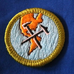 May include: A round, embroidered Boy Scout merit badge with a yellow border. The center of the badge is light blue with an orange silhouette of a mountain range and two crossed ice axes.