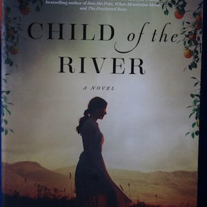 May include: Book cover with a woman walking in a field with a sunset in the background. The title of the book is "Child of the River" and the author is "Irma Joubert". The book is a novel and the author is also the author of "The Girl From The Train".