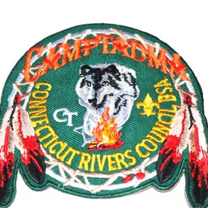 May include: Embroidered patch with a green background, featuring a wolf howling at a campfire, surrounded by a wreath of branches. The text "Camp Tad-ma Connecticut Rivers Council BSA CT" is written in a circle around the wolf. Two red and white feathers are on the bottom.