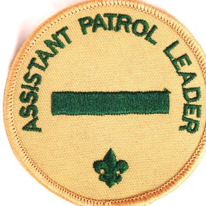 May include: A round, tan-colored patch with green embroidered text that reads "Assistant Patrol Leader". A green bar is centered on the patch, and a green fleur-de-lis is at the bottom.