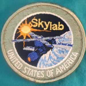 May include: A round, green embroidered patch with the words "Skylab" and "United States of America" in white. The patch features a blue and white space station orbiting a blue and white planet with a yellow sun in the background.