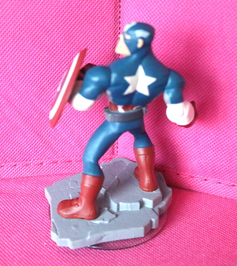 Puede incluir: Una figura de acci&oacute;n de Capit&aacute;n Am&eacute;rica azul y roja con una estrella blanca en el pecho, sosteniendo un escudo rojo, blanco y azul. La figura est&aacute; de pie sobre una base gris con un disco de pl&aacute;stico transparente debajo.