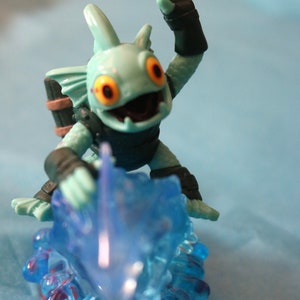 May include: A blue and green plastic toy figure of a creature with fins and a large mouth, riding a blue wave. The figure has orange eyes and is wearing a black and brown diving suit.