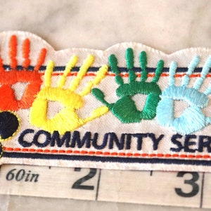 May include: Embroidered patch with a fleur-de-lis and rainbow handprints. The patch reads "COMMUNITY SERVICE" and measures approximately 4 inches in length.