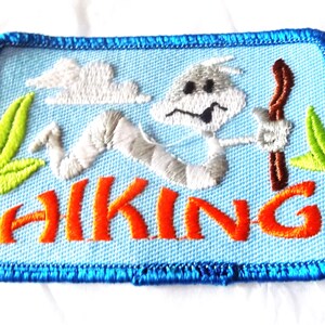 May include: A rectangular embroidered patch with a light blue background and a dark blue border. It features a white cartoon worm character holding a brown walking stick, white clouds, and green leaves. The word HIKING is stitched in orange.