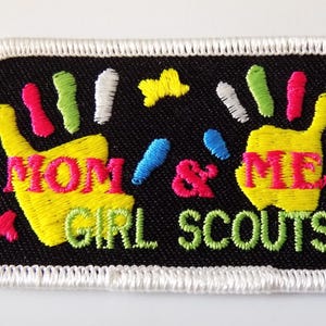 K&ouml;nnte beinhalten: Gestickter Aufn&auml;her mit schwarzem Hintergrund und wei&szlig;em, gen&auml;htem Rand. Der Aufn&auml;her zeigt zwei gelbe Handabdr&uuml;cke mit den Worten "MOM & ME" in Rosa und "GIRL SCOUTS" in Gr&uuml;n. Farbige Akzente sind Rosa, Blau, Gr&uuml;n und Gelb.