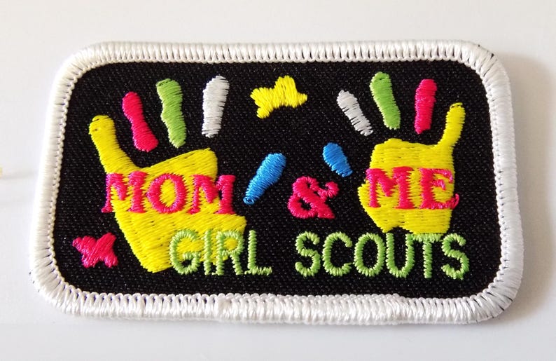 K&ouml;nnte beinhalten: Gestickter Aufn&auml;her mit schwarzem Hintergrund und wei&szlig;em, gen&auml;htem Rand. Der Aufn&auml;her zeigt zwei gelbe Handabdr&uuml;cke mit den Worten "MOM & ME" in Rosa und "GIRL SCOUTS" in Gr&uuml;n. Farbige Akzente sind Rosa, Blau, Gr&uuml;n und Gelb.
