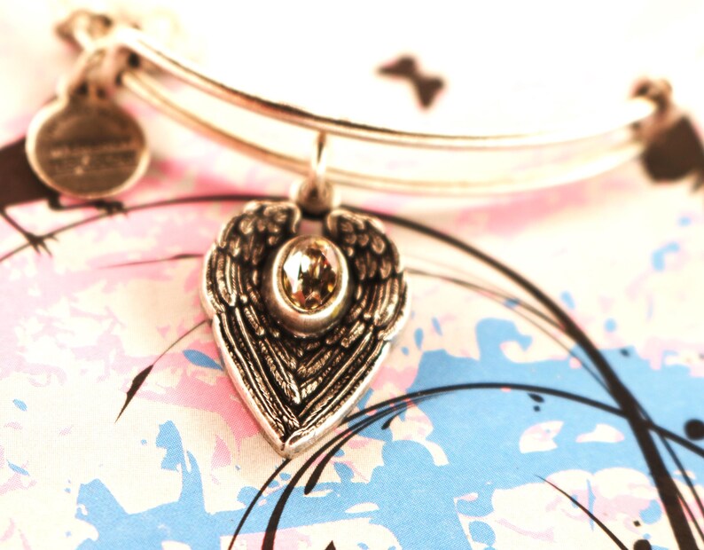 May include: A silver charm bracelet with a heart-shaped charm featuring a pair of angel wings and a clear crystal in the centre.