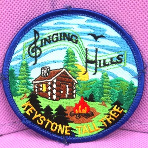 May include: A blue and green embroidered patch with a cabin, trees, a campfire, and the text "Singing Hills Keystone Tall Tree".
