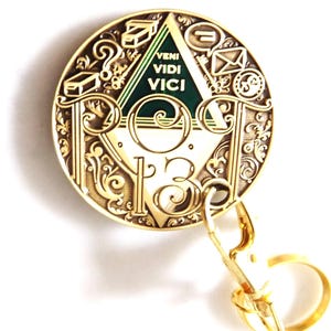 May include: Gold-colored round keychain with intricate scrollwork and the words "VENI VIDI VICI" in a green triangle. The keychain has a clasp and a ring, and the design includes symbols and the number 13.