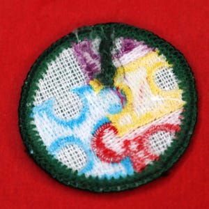 Pode incluir: Um remendo bordado circular de cor verde, branco, azul, amarelo e vermelho. O remendo tem um fundo branco com um desenho azul, amarelo e vermelho no centro. O remendo &eacute; rodeado por uma borda verde.