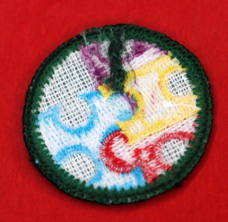 Pode incluir: Um remendo bordado circular de cor verde, branco, azul, amarelo e vermelho. O remendo tem um fundo branco com um desenho azul, amarelo e vermelho no centro. O remendo &eacute; rodeado por uma borda verde.