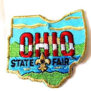 May include: Embroidered patch in the shape of Ohio with the text "OHIO STATE FAIR". The patch features a blue sky, green grass, and a gold fleur-de-lis. The word "OHIO" is in red and white.