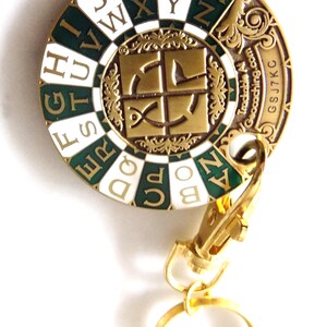 May include: A round, gold-colored metal keychain with a green and white enamel design. The design features letters of the alphabet and a central emblem. The keychain includes a clasp and a ring.