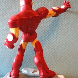 May include: Red and yellow Iron Man action figure with a raised arm and a white base. The figure is standing on a gray base with a circular design.