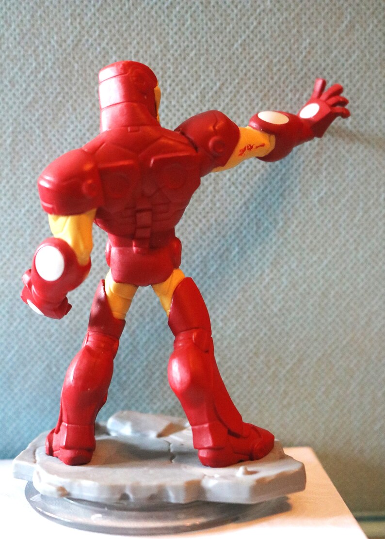 May include: Red and yellow Iron Man action figure with a raised arm and a white base. The figure is standing on a gray base with a circular design.