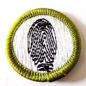 May include: Embroidered patch featuring a black fingerprint design on a white background. The circular patch is framed by a bright green stitched border. The design is a detailed representation of a fingerprint, suitable for clothing or accessories.