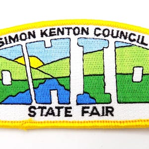 May include: Embroidered patch with a yellow border featuring the text "SIMON KENTON COUNCIL OHIO STATE FAIR 1997". The patch has a landscape design with green, blue, and yellow colors, and a black fleur-de-lis.