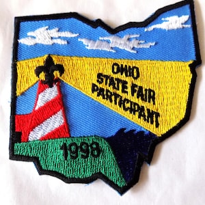 May include: Embroidered patch in the shape of Ohio, featuring a lighthouse and text. The patch has a blue sky with white clouds, a yellow field, and a green base. Text reads "OHIO STATE FAIR PARTICIPANT 1998".