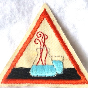 May include: A triangular shaped patch with an orange border. The center of the patch features a black background with a blue beaker and red fumes coming from the top.