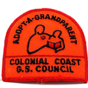 May include: An orange patch with black text and a cartoon bear reading a book. The text reads "ADOPT-A-GRANDPARENT" and "COLONIAL COAST G.S. COUNCIL". The patch has a red border.