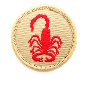 May include: A circular patch featuring a red scorpion embroidered on a tan background. The patch has a tan border with a stitched edge. The scorpion is detailed with multiple legs and a curved tail, set against a neutral backdrop.
