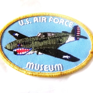 May include: Embroidered patch featuring a military aircraft with a shark mouth design. The patch has a light blue background with the text "U.S. AIR FORCE MUSEUM" and a gold border. The aircraft is green and black.