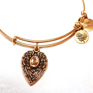May include: Gold charm bracelet with a heart-shaped charm featuring angel wings and a clear crystal. The bracelet has two additional charms, one with the text "I love" and the other with a small American flag.