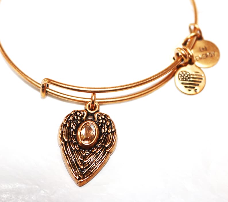 May include: Gold charm bracelet with a heart-shaped charm featuring angel wings and a clear crystal. The bracelet has two additional charms, one with the text "I love" and the other with a small American flag.