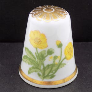 May include: White porcelain thimble with a gold rim and a gold floral design on the top. The thimble features a hand-painted design of yellow flowers with green leaves and buds.