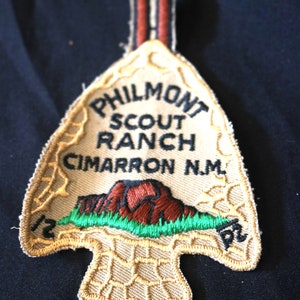 May include: A tan felt arrowhead patch with the text "Philmont Scout Ranch Cimarron N.M." in black embroidery. The patch features a red embroidered mountain range with green grass and a tan embroidered border.